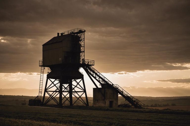 Photographie d'une structure de mine de charbon en silhouette contre un coucher de soleil qui s'estompe, avec la campagne bourguignonne en toile de fond, en tons sépia vintage, éclairage ambiant, mise au point nette.