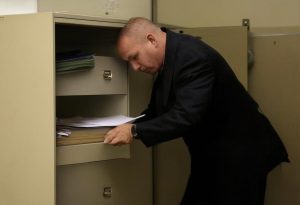 Un huissier examine prudemment un classeur ouvert dans un bureau encombré, suggérant une recherche de documents cachés.