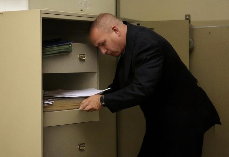 Un huissier examine prudemment un classeur ouvert dans un bureau encombré, suggérant une recherche de documents cachés.